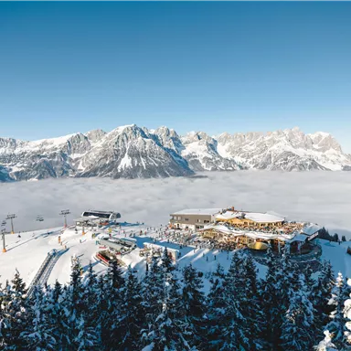 A beautiful winter landscape with snow-covered mountains and a clear sky. In the foreground, ski slopes and a mountain hut can be seen.