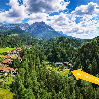 Eine malerische Landschaft mit Bergen und dichten Wäldern. Im Vordergrund sind bunte Häuser und eine Beschilderung zum Talschuster zu sehen.