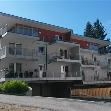 Ein modernes Wohngebäude mit mehreren Balkonen und großen Fenstern. Die Umgebung ist ruhig mit Bäumen im Hintergrund.