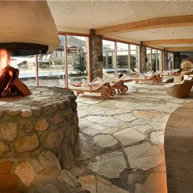 A cozy interior with a stone fireplace and a fire pit. In the background are loungers and large windows overlooking nature.
