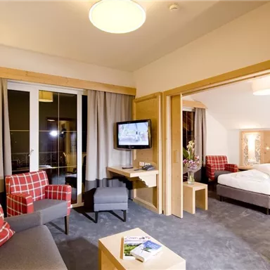 Ein modernes Hotelzimmer mit einem Wohnbereich und einem Schlafzimmer. Die Einrichtung ist gemütlich, mit einem Fernseher und großen Fenstern für viel Licht.