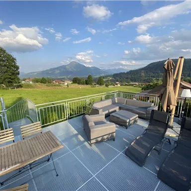 A cozy terrace with seating and loungers. In the background, green meadows and mountains can be seen.