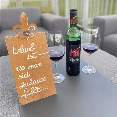 A stylish arrangement with a bottle of red wine and two glasses on a table. Next to the wine bottle is a sign that reads "Vacation is where you feel at home."