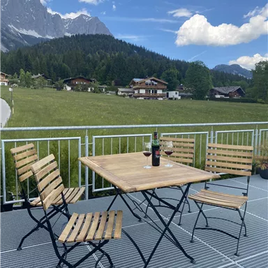 A cozy table with chairs on the terrace, surrounded by a beautiful landscape with mountains. A wine bottle and glasses are ready to enjoy the view.