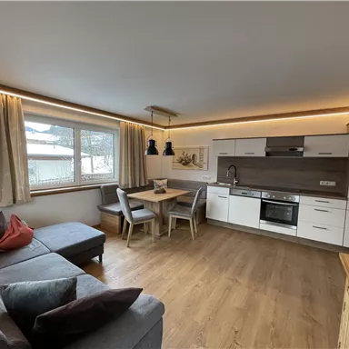 A modern living room with a cozy couch and a dining table. The bright kitchen is well-equipped and has large windows for plenty of daylight.