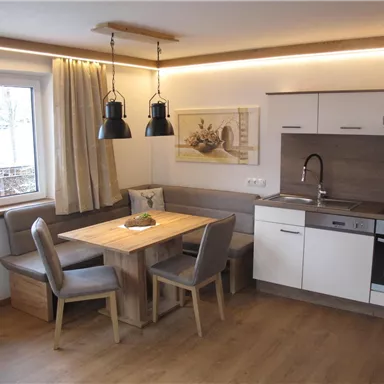 A modern kitchen with a dining table and seating area. The room has light-colored walls and wooden floors, with windows that let in natural light.
