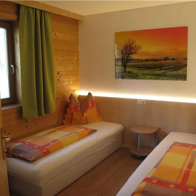 A cozy room with two single beds, colorful pillows, and wooden walls. A window lets in plenty of light, and a picture of a tree adorns the wall.