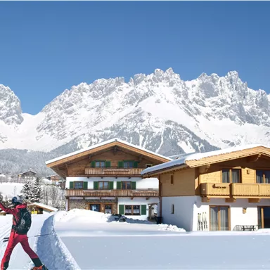 Two skiers are enjoying the snow in front of chalets in a winter mountain landscape. In the background, majestic mountains rise beneath a radiant blue sky.