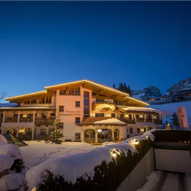 Ein charmantes Hotel im Schnee, beleuchtet von warmen Lichtern. Im Hintergrund sind schneebedeckte Berge und der Abendhimmel zu sehen.
