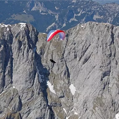 A paraglider glides over an impressive mountain landscape. The steep, gray rocks and snow-covered peaks create a spectacular backdrop.