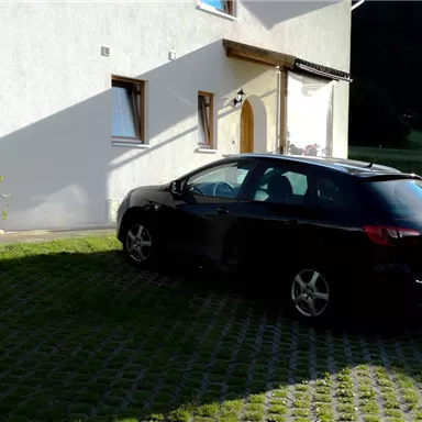 Ein schwarzes Auto parkt vor einem Haus. Die Umgebung ist grün mit einem gepflasterten Gehweg.