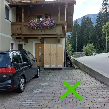Ein Parkplatz neben einem Holzhaus mit Balkon. Rechts ist ein grünes "X" auf dem Boden markiert.