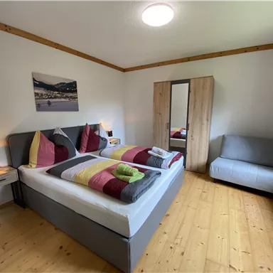 A cozy bedroom with a large bed, colorful pillows, and a sofa. The wooden floors and warm colors create an inviting atmosphere.