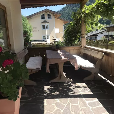 A cozy terrace with a long wooden table and benches. In the foreground are colorful flowers and the surroundings are surrounded by mountain landscapes.