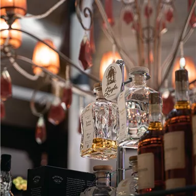 Eine elegante Bar mit beleuchteten Lampen und einer Auswahl an hochwertigen Spirituosen. Im Vordergrund stehen mehrere Glasflaschen mit verschiedenen Getränken.