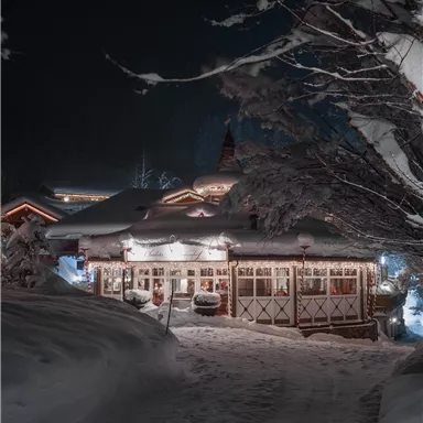 Ein gemütliches Gebäude im Schnee, beleuchtet von warmem Licht. Der Weg ist von frischem Schnee umgeben, und die Atmosphäre ist einladend und winterlich.