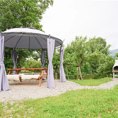 Ein gemütlicher Pavillon mit einer hängenden Bank und grauen Vorhängen in einer grünen Landschaft. Im Hintergrund steht ein Grill und einige Bäume.