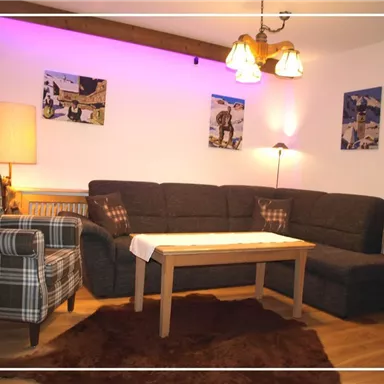 Ein gemütliches Wohnzimmer mit einer dunklen Couch und einem Holztisch. An den Wänden hängen Bilder und eine sanfte Beleuchtung schafft eine warme Atmosphäre.