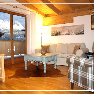 Ein gemütliches Wohnzimmer mit Holzdecke und warmen Farben. Im Hintergrund Sicht auf schneebedeckte Berge durch ein großes Fenster.