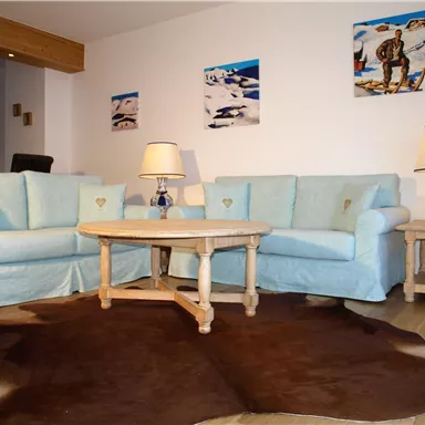 Ein gemütliches Wohnzimmer mit zwei hellblauen Sofas und einem Holztisch. An der Wand hängen farbige Bilder, und der Boden ist mit einem Fellteppich bedeckt.