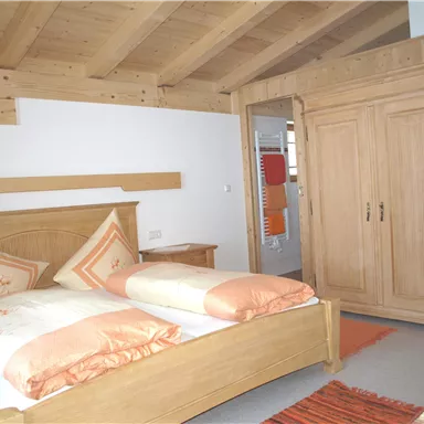 Ein gemütliches Zimmer mit einem Holzbett und orangefarbenen Dekokissen. Der Raum hat eine helle Holzdecke und einen großen Kleiderschrank.