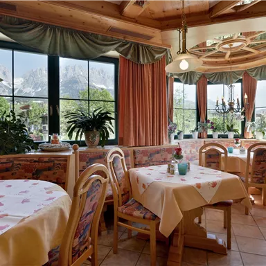 Ein gemütliches Restaurant mit Holzmöbeln und hellen Tischen. Große Fenster bieten einen Blick auf die Natur und die Berge.