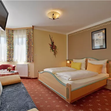 Ein gemütliches Schlafzimmer mit einem Doppelbett und bunten Kissen. Es gibt ein Sofa, einen Fernseher und große Fenster, die viel Licht hereinlassen.