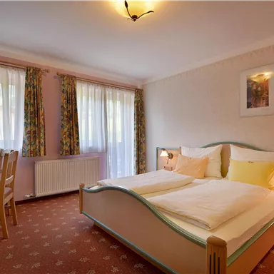 Ein gemütliches Schlafzimmer mit einem Doppelbett, einem Tisch und Stühlen. Große Fenster sorgen für viel Tageslicht und eine einladende Atmosphäre.