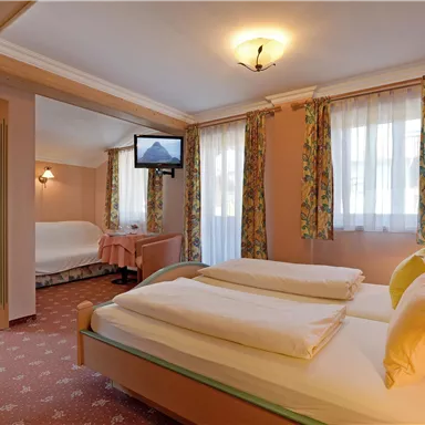 Ein gemütliches Hotelzimmer mit zwei Betten und Fernseher. Es gibt große Fenster mit Vorhängen und einen separaten Schlafbereich.