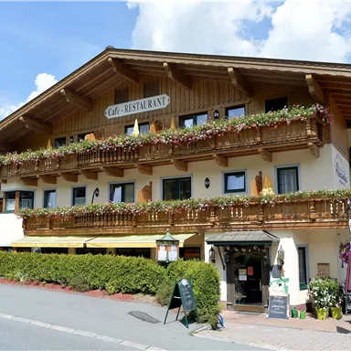Ein charmantes Gebäude mit einem Restaurant-Schild und vielen blühenden Balkonkästen. Der Himmel ist blau und es gibt eine grüne Umgebung.