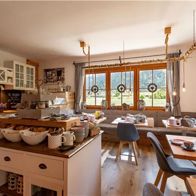 Ein gemütlicher Frühstücksraum mit Holzmöbeln und großen Fenstern. Auf dem Tisch sind verschiedene Speisen und Getränke angerichtet.