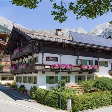 Ein charmantes alpinisches Haus mit bunten Blumenfenstern und Solarzellen. Umgeben von grüner Vegetation und Bergen im Hintergrund.