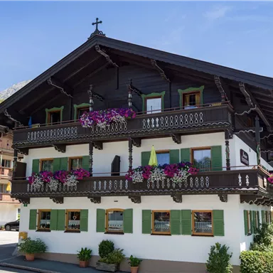 Ein traditionelles alpenländisches Holzhaus mit grünen Fensterläden und bunten Blumen auf dem Balkon. Im Hintergrund sind die Berge und ein klarer blauer Himmel sichtbar.