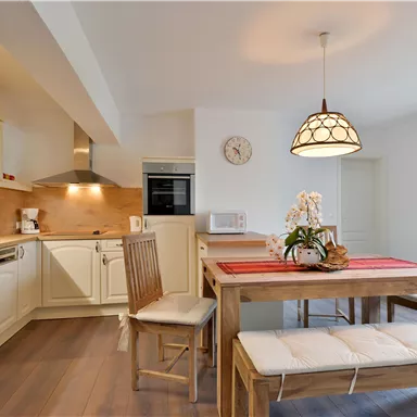 Eine helle Küche mit modernen Möbeln und einem Esstisch aus Holz. Die Wände sind neutral und es gibt farbenfrohe Vorhänge an einem Fenster.