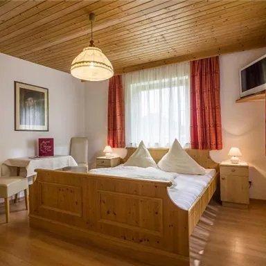 Ein gemütliches Schlafzimmer mit einem Holzbett und zwei Kissen. Die Wände sind hell, und der Raum hat eine warme Atmosphäre mit Fenstern und Vorhängen.