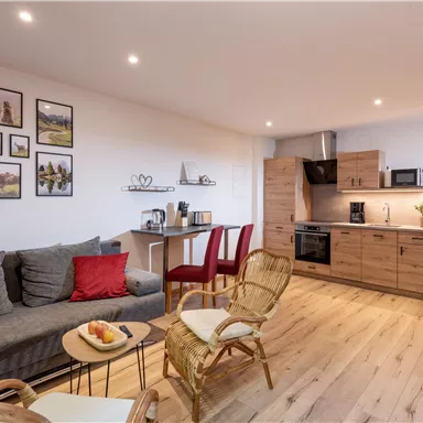 Ein modernes Wohnzimmer mit einem gemütlichen Sofa und einem Esstisch. Die Einrichtung ist hell und einladend, mit Bildern an der Wand und einer Küchenzeile.