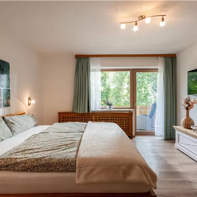 Ein helles Schlafzimmer mit einem großen Bett und warmen Farbtönen. Durch das Fenster ist ein Blick ins Grüne möglich.