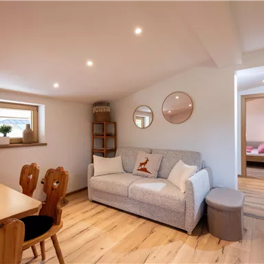 Ein helles, modernes Wohnzimmer mit einem gemütlichen Sofa und einem Esstisch. Der Raum hat große Fenster und freundliche, neutrale Farben.