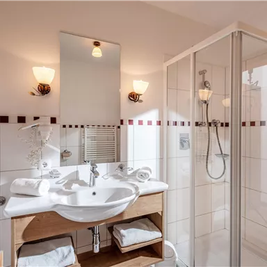 Ein modernes Badezimmer mit einer Dusche und einem Waschbecken. Die Wände sind hell gefliest und es gibt Handtücher auf einem Regal.