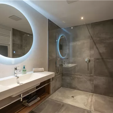 Ein modernes Badezimmer mit einem großen Spiegel und einer begehbaren Dusche. Die Wände sind in einem schlichten Grauton gehalten, mit eleganten Armaturen und Handtüchern.