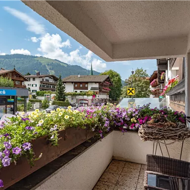 Ein Balkon mit bunten Blumen und Blick auf die Berge. Im Hintergrund sind Häuser und ein klarer Himmel zu sehen.