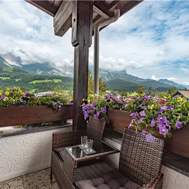 Ein gemütlicher Balkon mit einem Tisch und Stühlen, umgeben von bunten Blumen. Im Hintergrund sind beeindruckende Berge und ein klarer Himmel zu sehen.