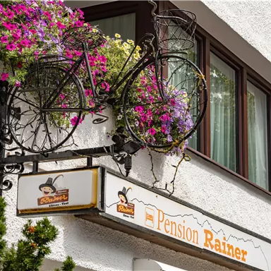 Eine charmante Pension mit einem schmiedeeisernen Fahrrad und bunten Blumen im Fenster. Das Schild zeigt den Namen "Pension Rainer".