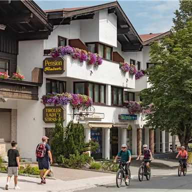 Eine malerische Straße mit traditionellen Gebäuden und bunten Blumenkästen. Menschen gehen und fahren mit dem Fahrrad vorbei, während Bäume Schatten spenden.