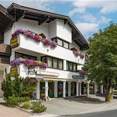 Ein traditionelles Gebäude mit Blumenkästen und Bäumen vor der Fassade. Der Himmel ist teilweise bewölkt und die Umgebung wirkt einladend.