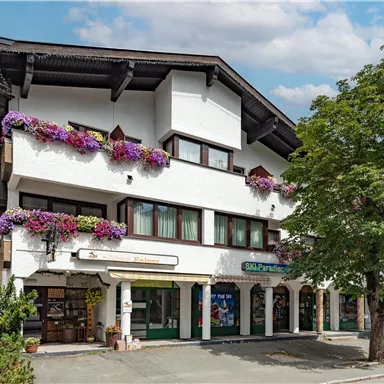 Ein typisch alpenländisches Gebäude mit blühenden Balkonen. Die Fassade ist hell und freundlich, umgeben von Bäumen.