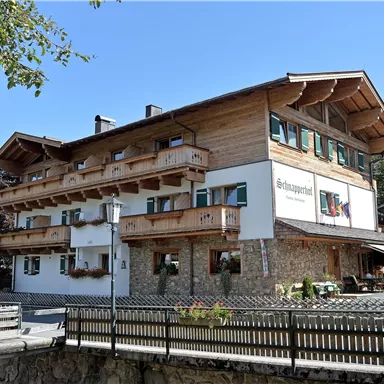 Ein traditionelles alpenländisches Gebäude mit Balkon und Holzverkleidung. Umgeben von Bäumen und einem klaren blauen Himmel.