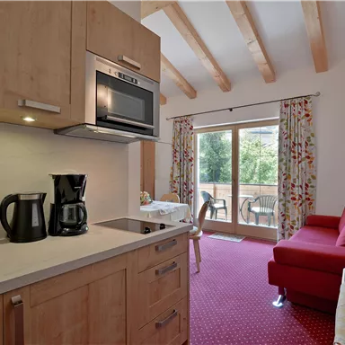 Eine moderne Küchenzeile mit Kochfeld, Mikrowelle und Kaffeemaschine. Im Hintergrund befindet sich ein gemütlicher Wohnbereich mit rotem Sofa und großen Fenstern.