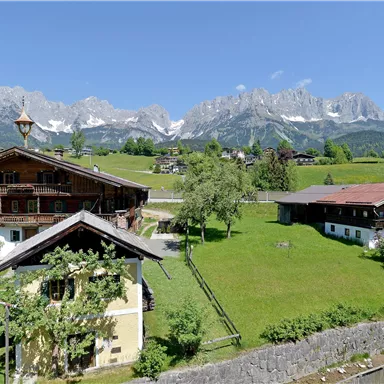 Eine idyllische Alpenlandschaft mit traditionellen Holzhäusern. Im Hintergrund sind majestätische Berge und ein klarer blauer Himmel zu sehen.