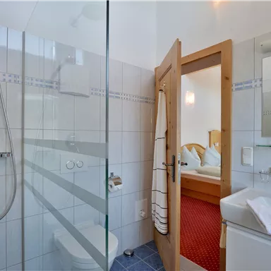 Ein modernes Badezimmer mit einer Glasdusche und einem Waschbecken. Hinter der Dusche ist ein heller Schlafbereich sichtbar.